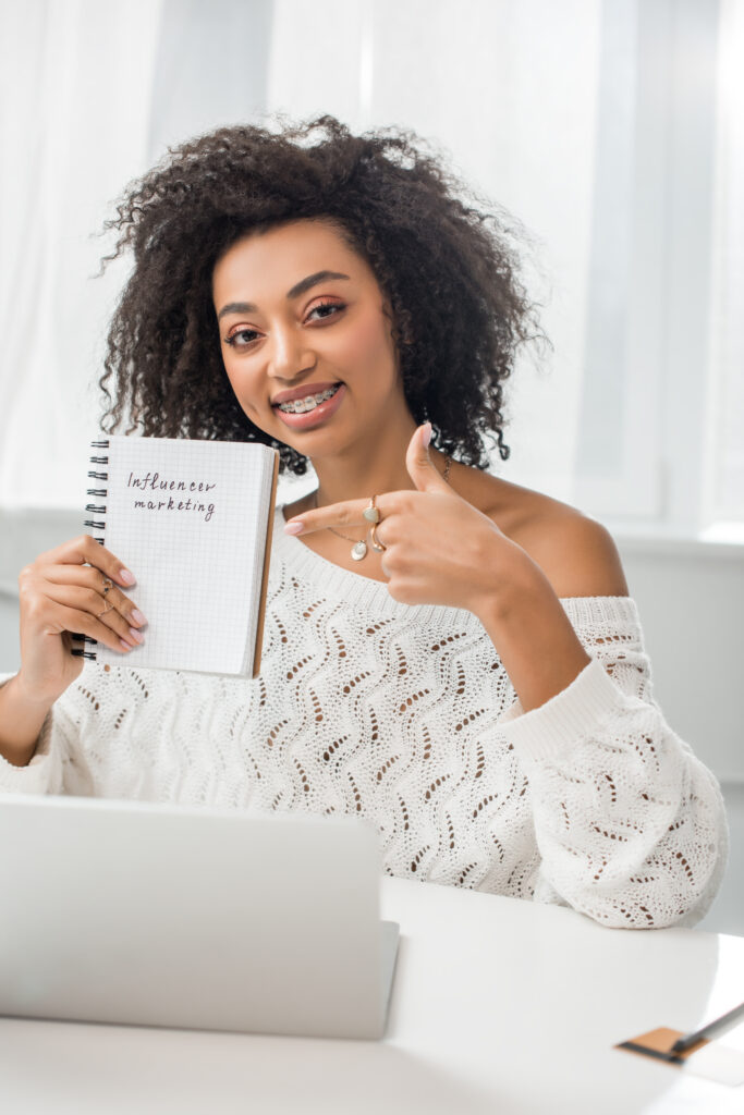 cheerful african american girl in braces pointing with finger at notebook with influencer marketing lettering at home
