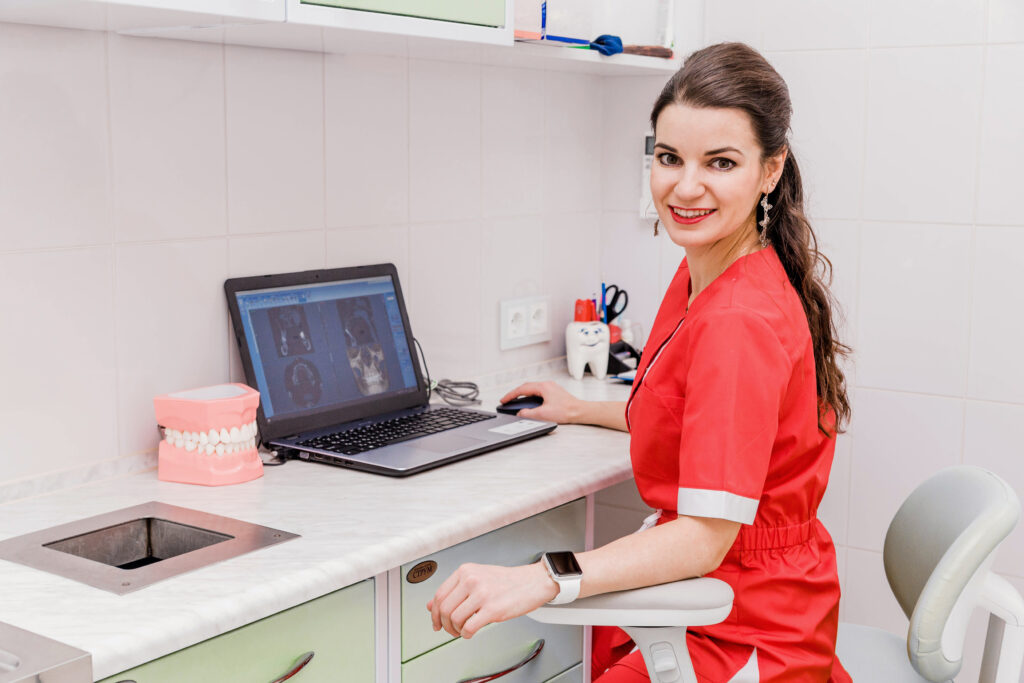 An attractive young female dentist sits at a table and works on a laptop where the results of the examination are visible.